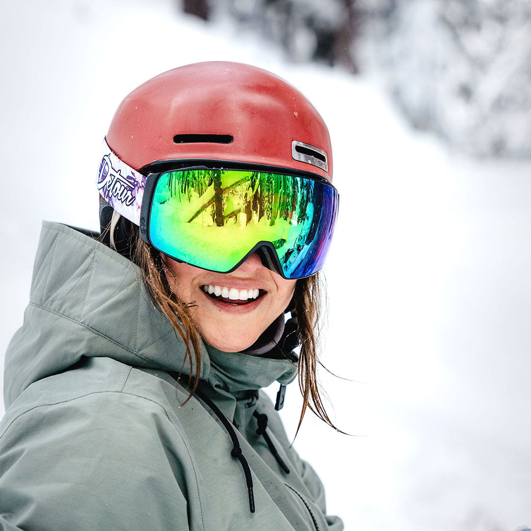 Skier in colorful goggles and patterned jacket holding skis over shoulder in snowy landscape.